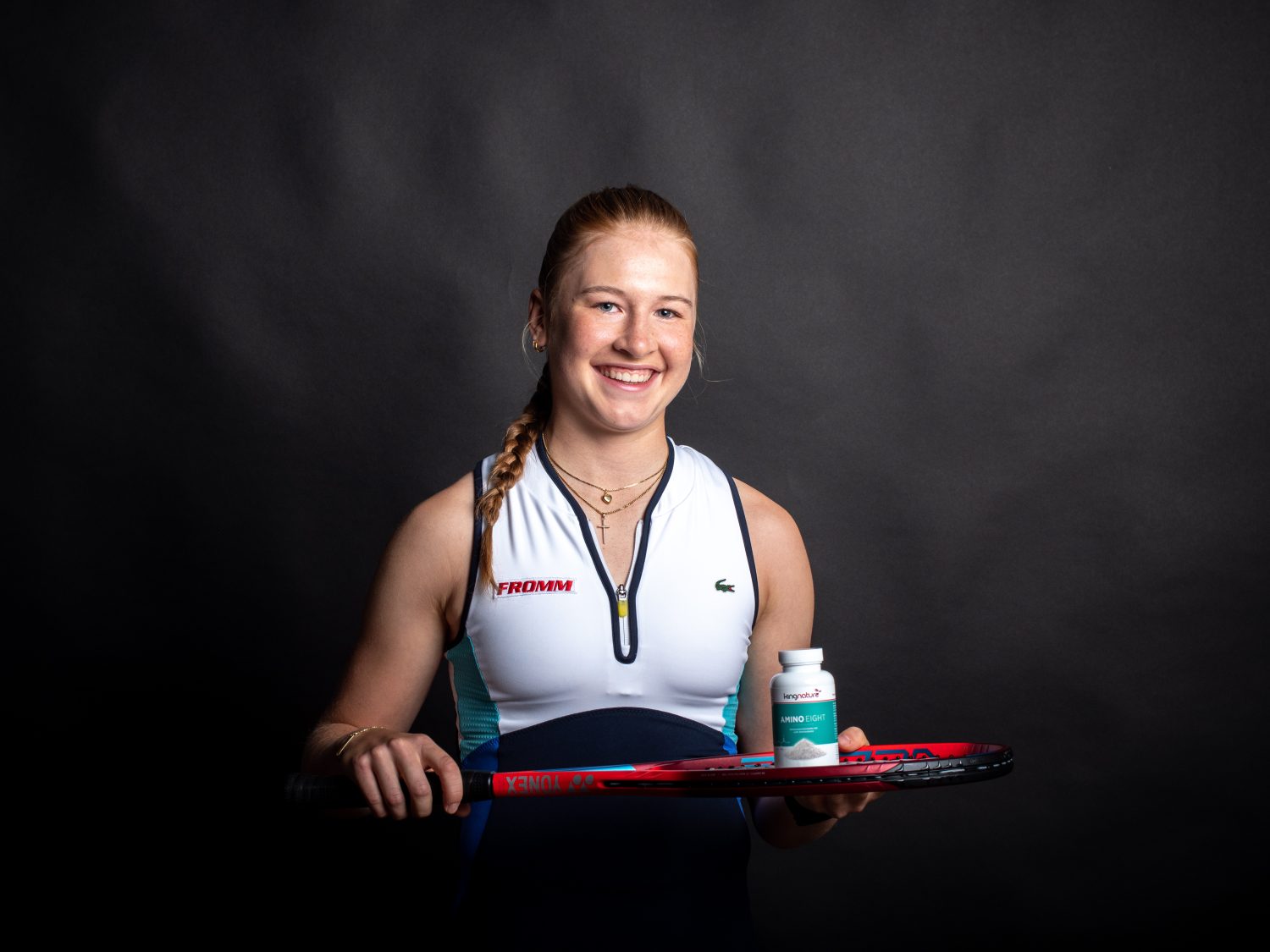 A smiling woman in athletic wear holds a tennis racket and a bottle of supplements against a dark background.