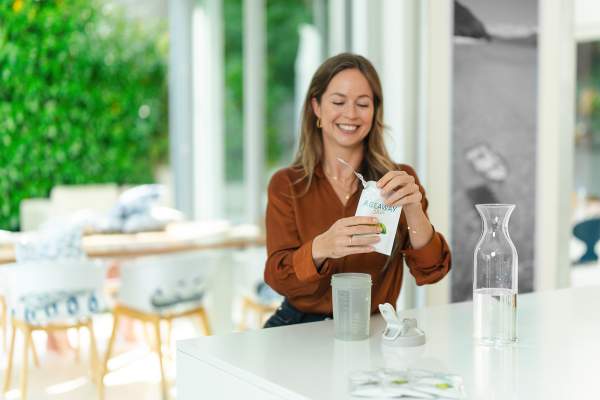 Eine lächelnde Frau sitzt an einer hellen Küchentheke und öffnet eine Packung „AG1“ von kingnature Nahrungsergänzung aus der Schweiz neben einer Glaskaraffe mit Wasser, einer Shaker-Flasche und anderen Packungen mit Nahrungsergänzungsmitteln.