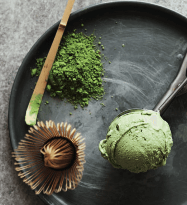 Auf einem schwarzen Teller liegen eine Kugel grünes Matcha-Eis, ein Bambusbesen, ein Löffel und Matcha-Pulver – ideal für alle, die Matcha Pulver Bio kunstvoll arrangiert kaufen möchten.
